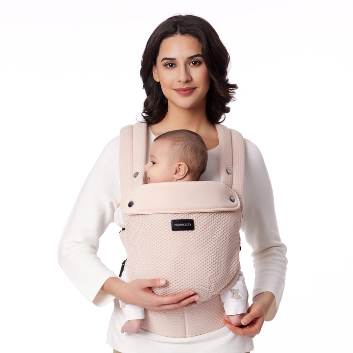 Woman in beige holding baby in Air Mesh-Pink Momcozy ergonomic carrier, promoting comfort.