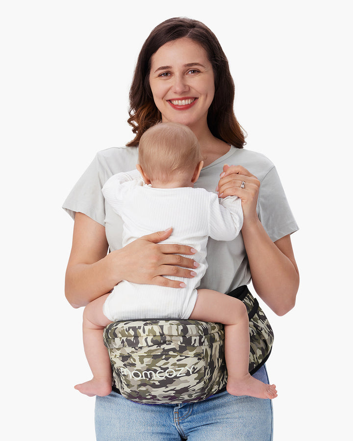 Woman using a camouflage-patterned Momcozy baby hip seat carrier holding a baby in a white onesie, showcasing the 3D Belly Protector & EVA Massage Board feature for added comfort.