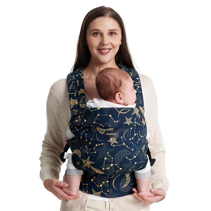 Woman holding a newborn using a Momcozy baby carrier with Starry Night design, dark blue background with golden stars and constellations.